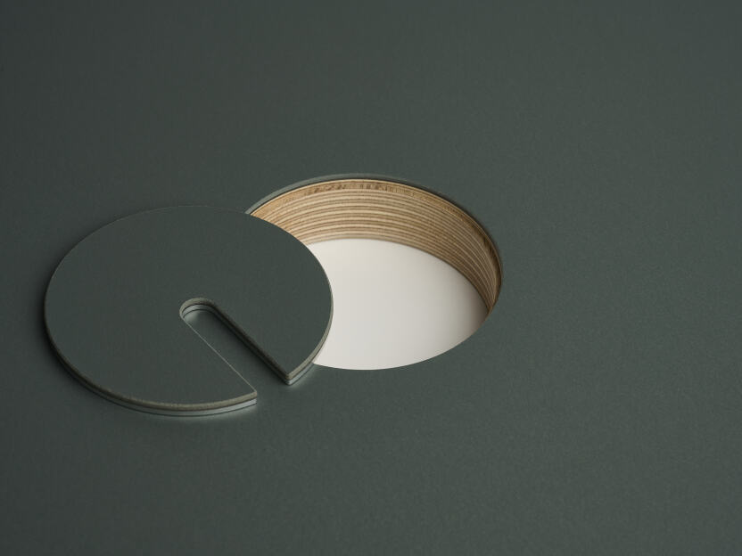 Eine eloxierte Aluminium-Rundabdeckung für Kabel von Daniel Lorch, die auf einer Seite mit Conifer-Linoleum bedeckt ist. Sie liegt neben der Aussparung der Tischplatte und offenbart das Sperrholz der Tischplattenkernmaterialien.