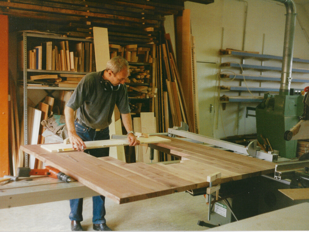 Person schneidet in einer Holzwerkstatt eine Tischplatte zu, umgeben von Werkzeugen, Maschinen und Regalen mit Holzmaterialien.