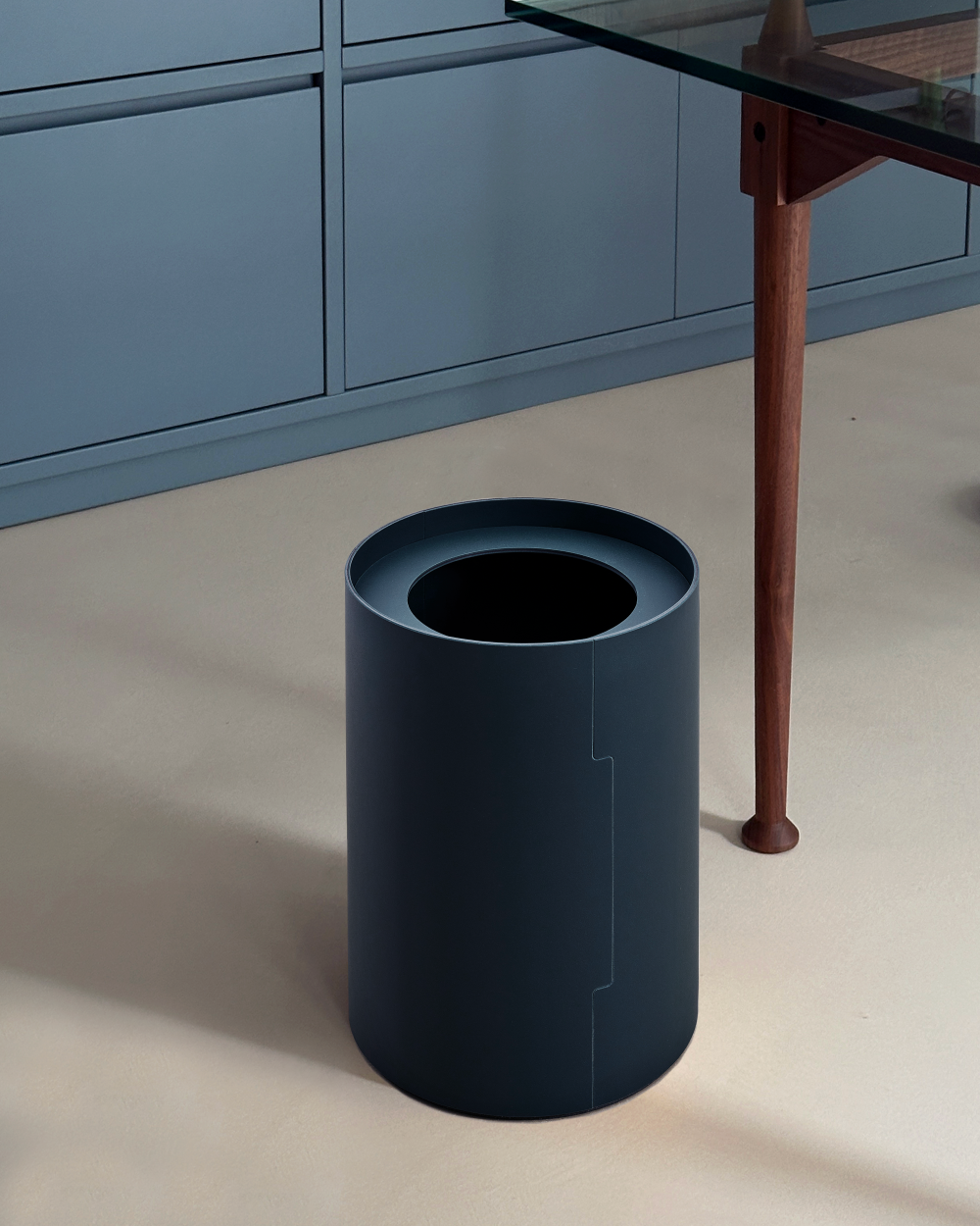 Cylindrical wastepaper basket in denim blue next to a glass table with wooden legs, in front of a blue cupboard.