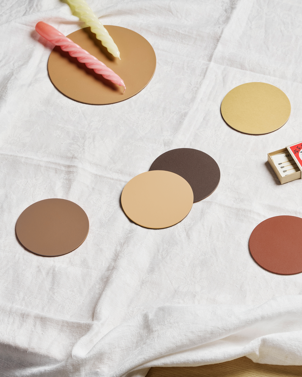 PAD coasters made of linoleum in warm colours arranged on a white tablecloth, with coloured candles and a matchbox.