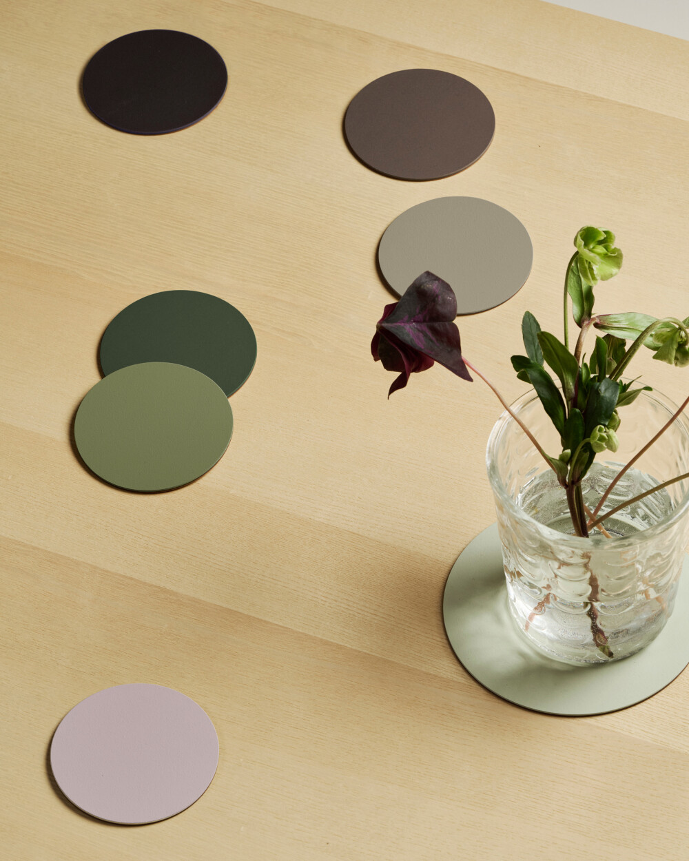 PAD coasters made of linoleum in various colours (Wildwood collection), arranged on a light wooden table with a glass vase.