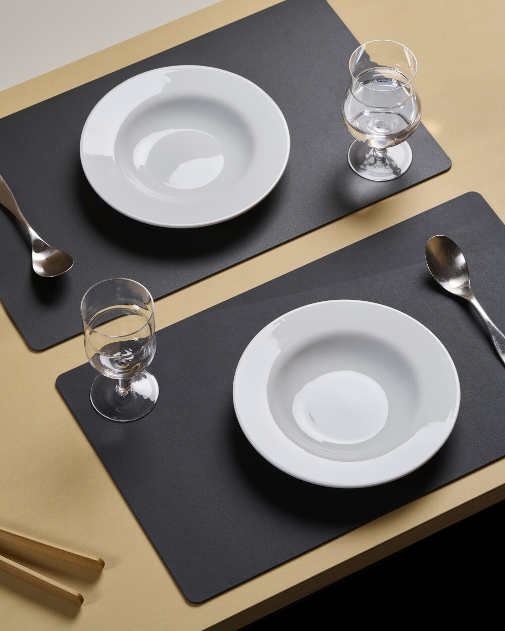 Two PAD placemats made of charcoal-coloured linoleum, covered with white plates, glasses and cutlery on a light-coloured table.